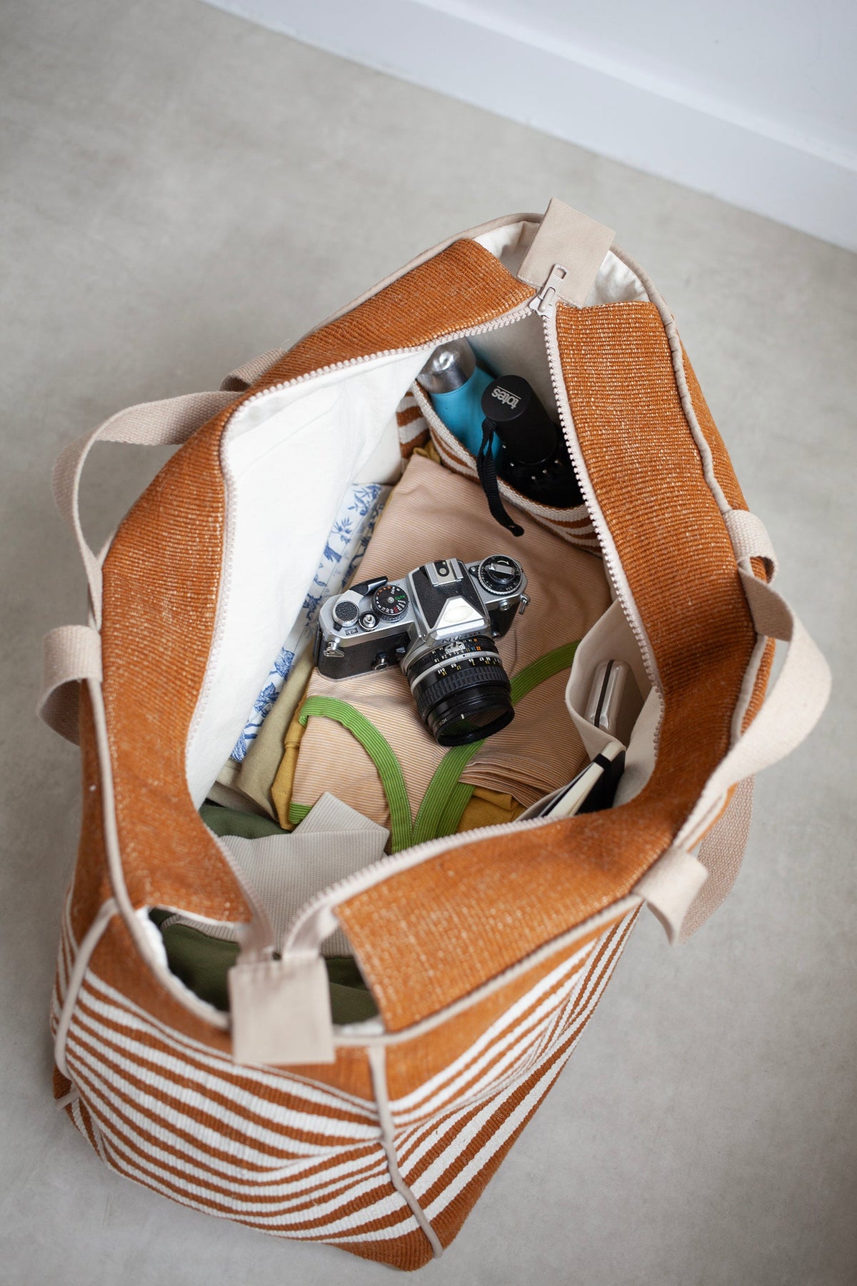 Inside view of brown and beige striped bag with camera and items on a light grey floor.
