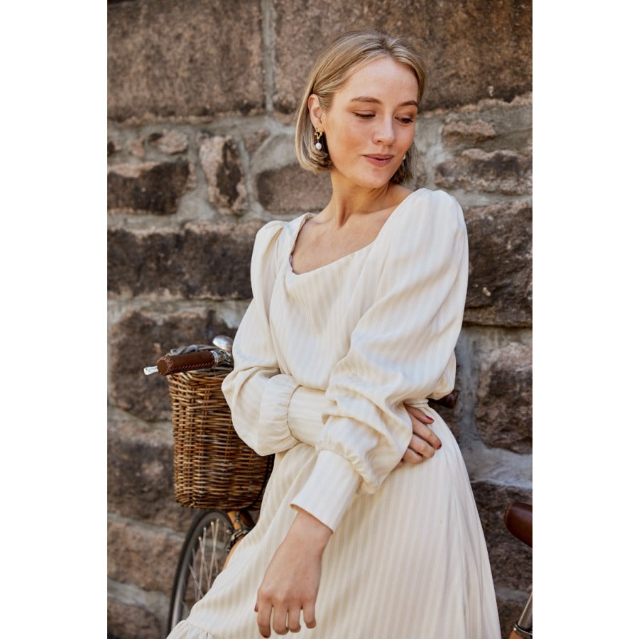 Close up of a white woman with short blonde hair standing against a brick wall and wearing a long sleeved cream dress