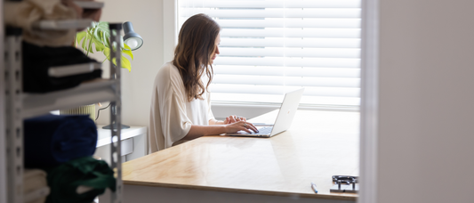 Woman sitting working on laptop at a large wooden table in front of window with bolts of fabric in the foreground