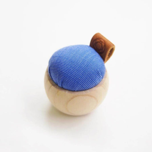 Small pincushion with pale wooden base, blue cushion and leather tag against white background