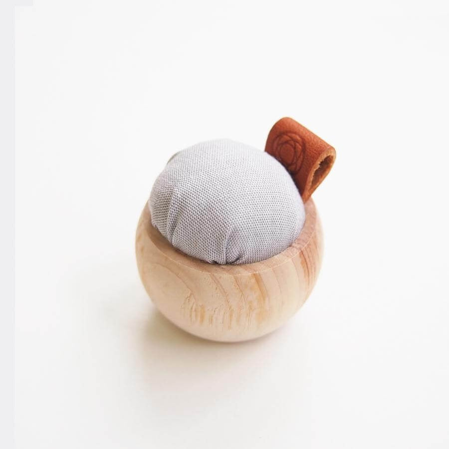 Small pincushion with pale wood base, grey fabric and leather tag against white background