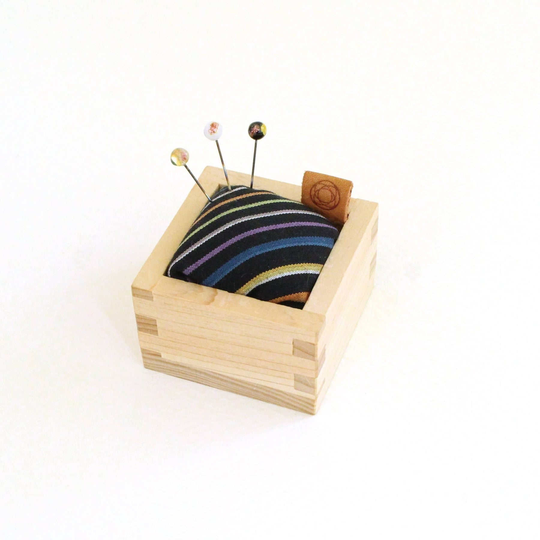 Square pin cushion with wooden base, black and colourful striped fabric with three pins sticking out