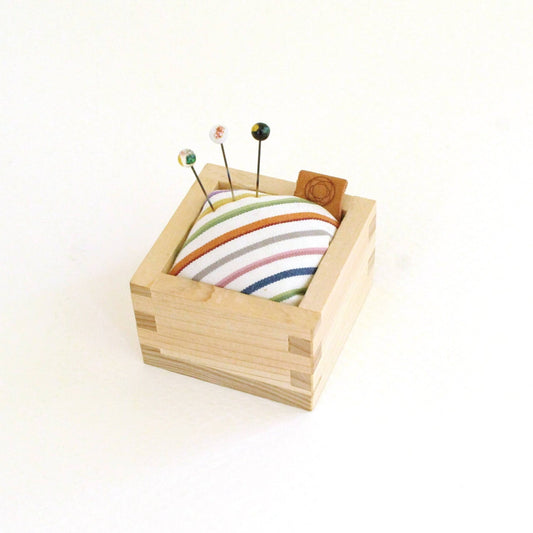Square cohana pin cushion with wooden base and white and colourful striped fabric with three pins sticking out
