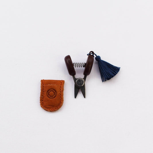 Mini scissors with blue tassel alongside leather case against white background