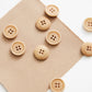 Natural light wood buttons with four holes scattered on a textured beige fabric square against a white background.