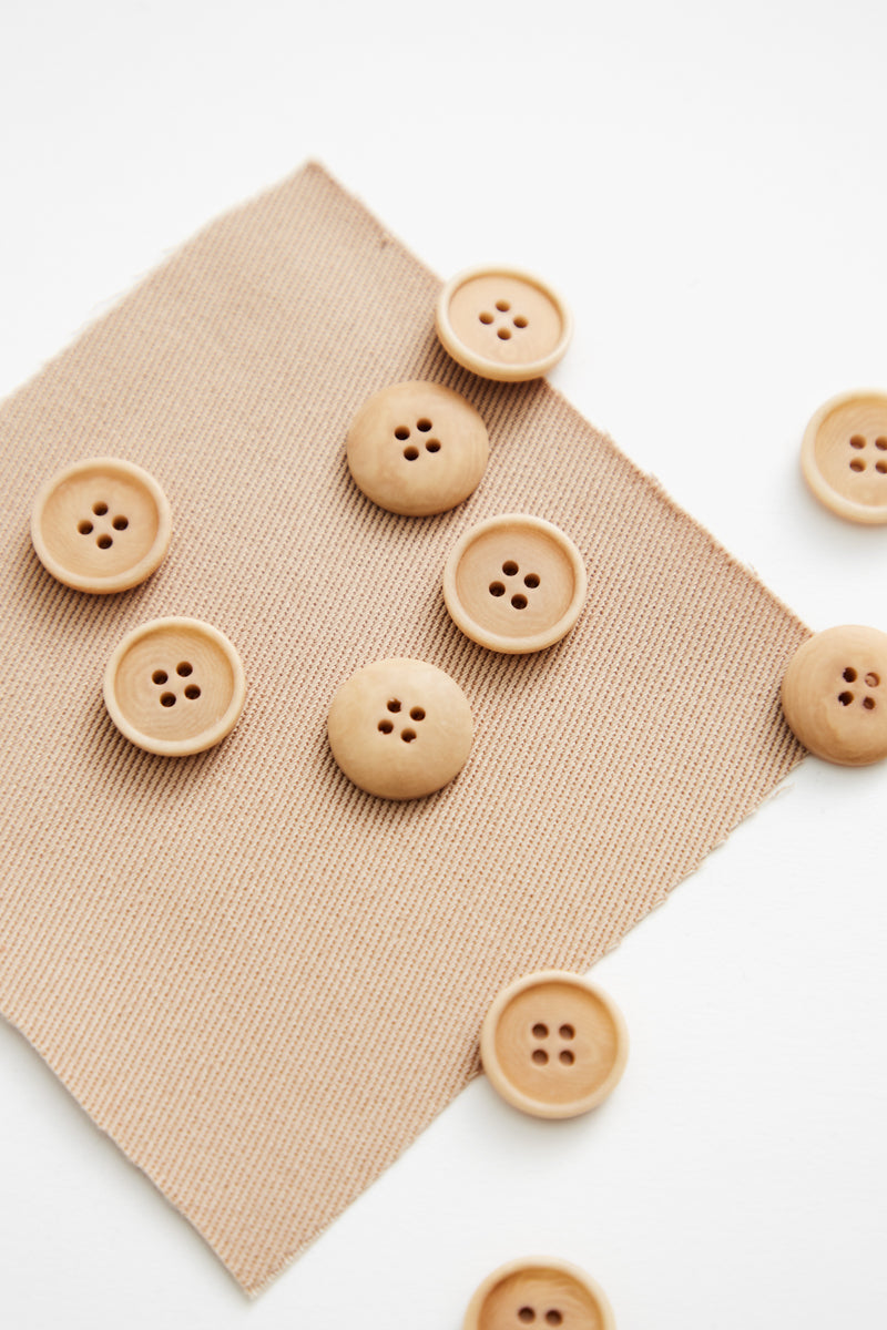 Natural light wood buttons with four holes scattered on a textured beige fabric square against a white background.