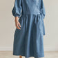 A woman wearing a blue wrap-style midi dress with three-quarter sleeves and a tie waist, standing barefoot in a neutral indoor setting.