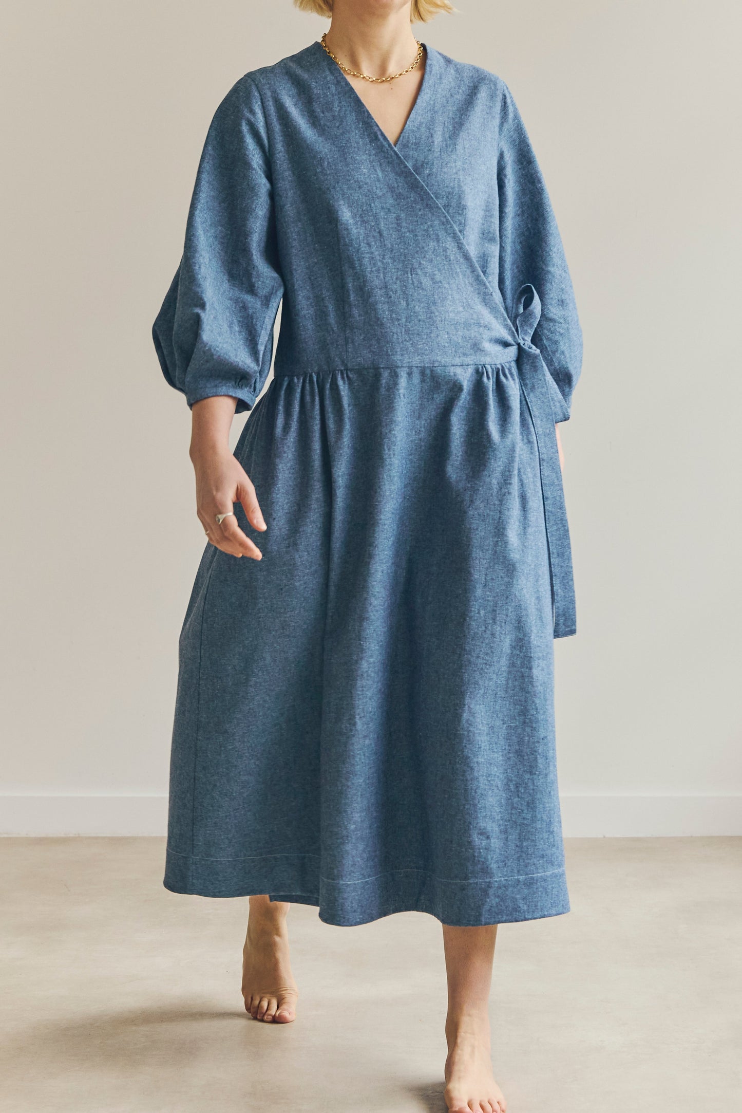 A woman wearing a blue wrap-style midi dress with three-quarter sleeves and a tie waist, standing barefoot in a neutral indoor setting.