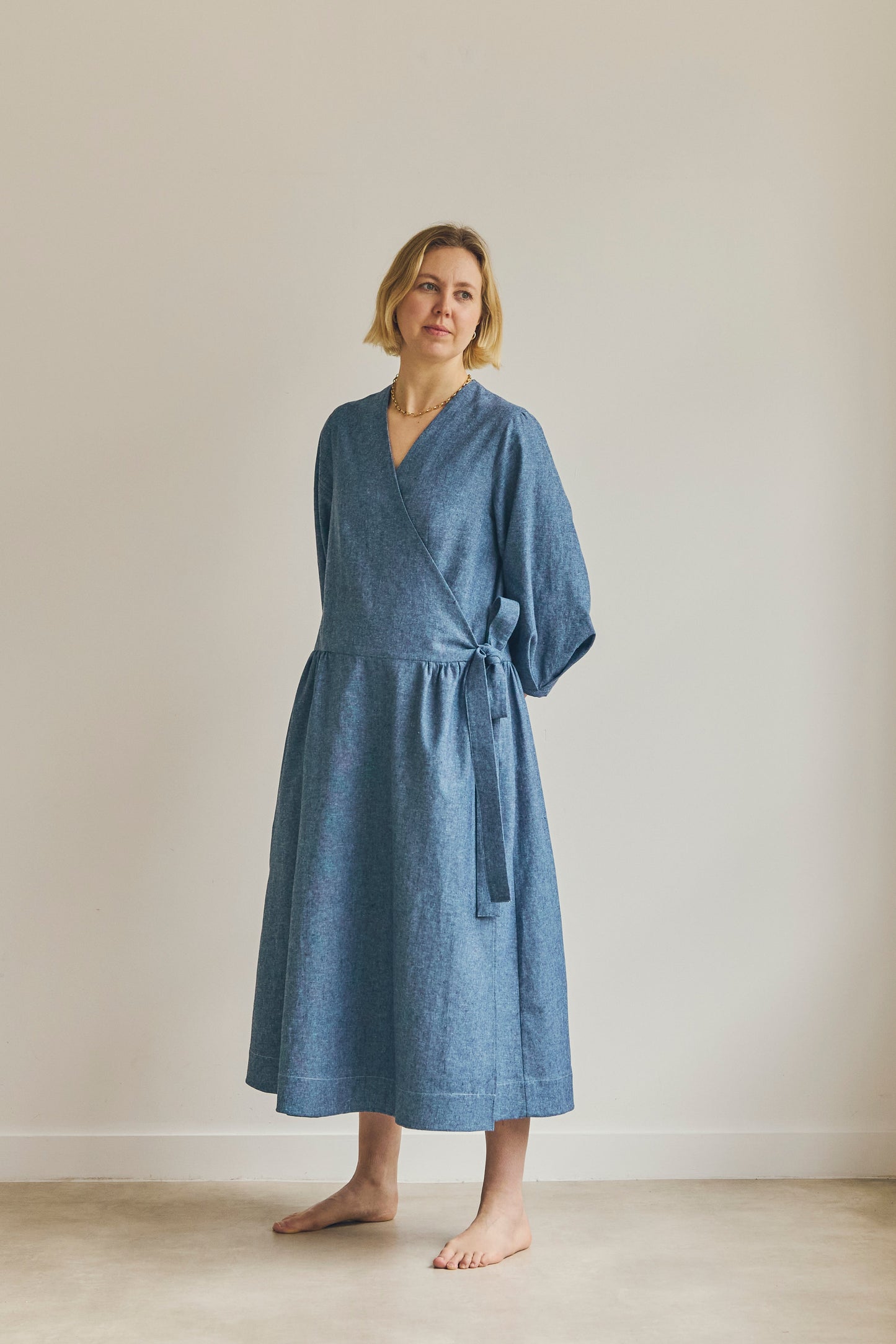 A woman wearing a blue linen wrap dress with a tie waist, standing barefoot in a minimalist studio setting with a beige wall and floor.