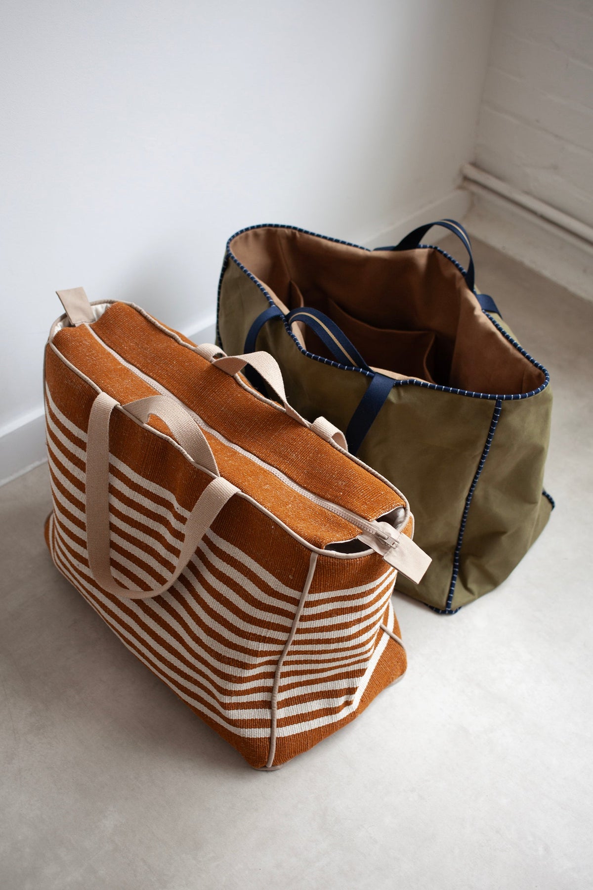 Two tote bags, one brown with closed zipper and white stripes, and one green, on a light floor.