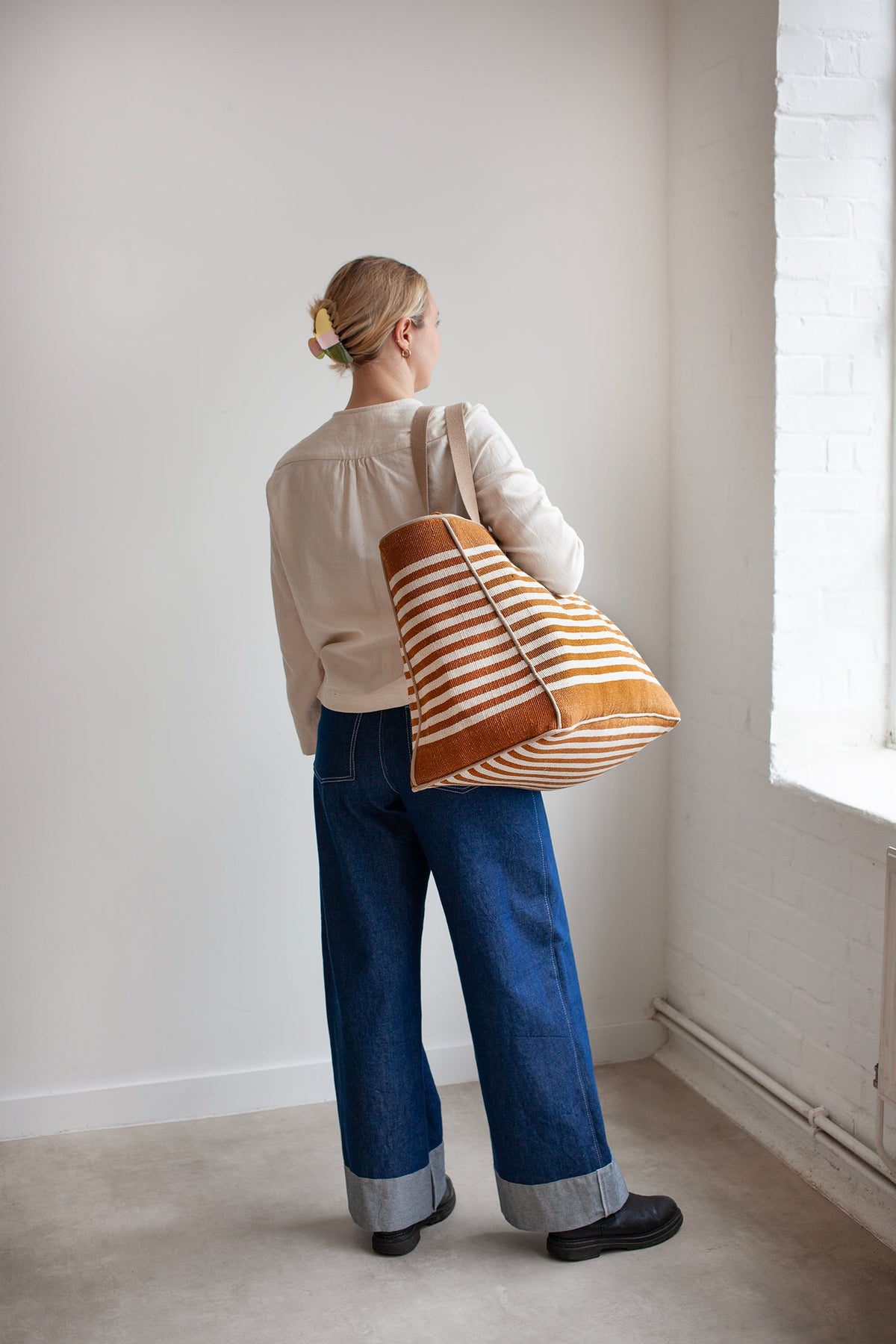 Person holding a striped Away tote bag in a minimalistic room with a window.