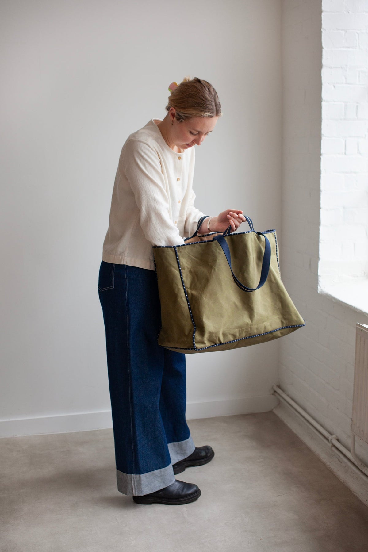 Person holding a green Away tote bag in a minimalistic room with white walls and a radiator.