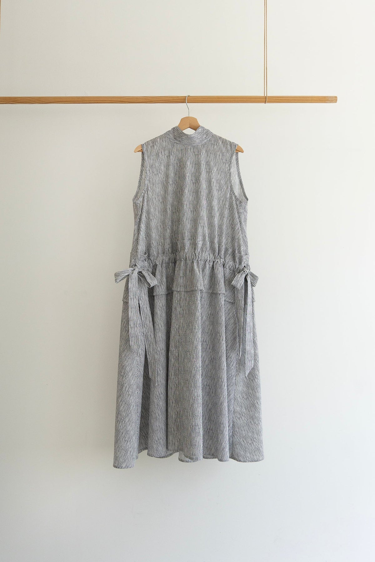 A sleeveless grey and white dress with side tie details, hanging on a wooden hanger from a wooden clothing rack against a plain white wall.