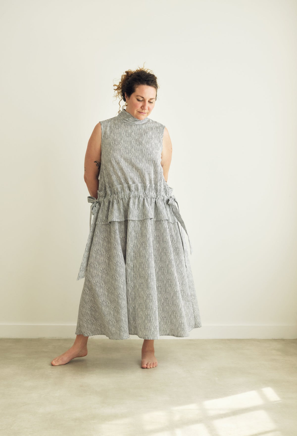 A woman stands barefoot in a minimalist room wearing a sleeveless, high-neck, midi dress with a grey and white pattern.