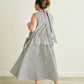 A woman wearing a sleeveless grey and white striped dress with a tie-back neckline, shown from behind in a minimalist studio setting.