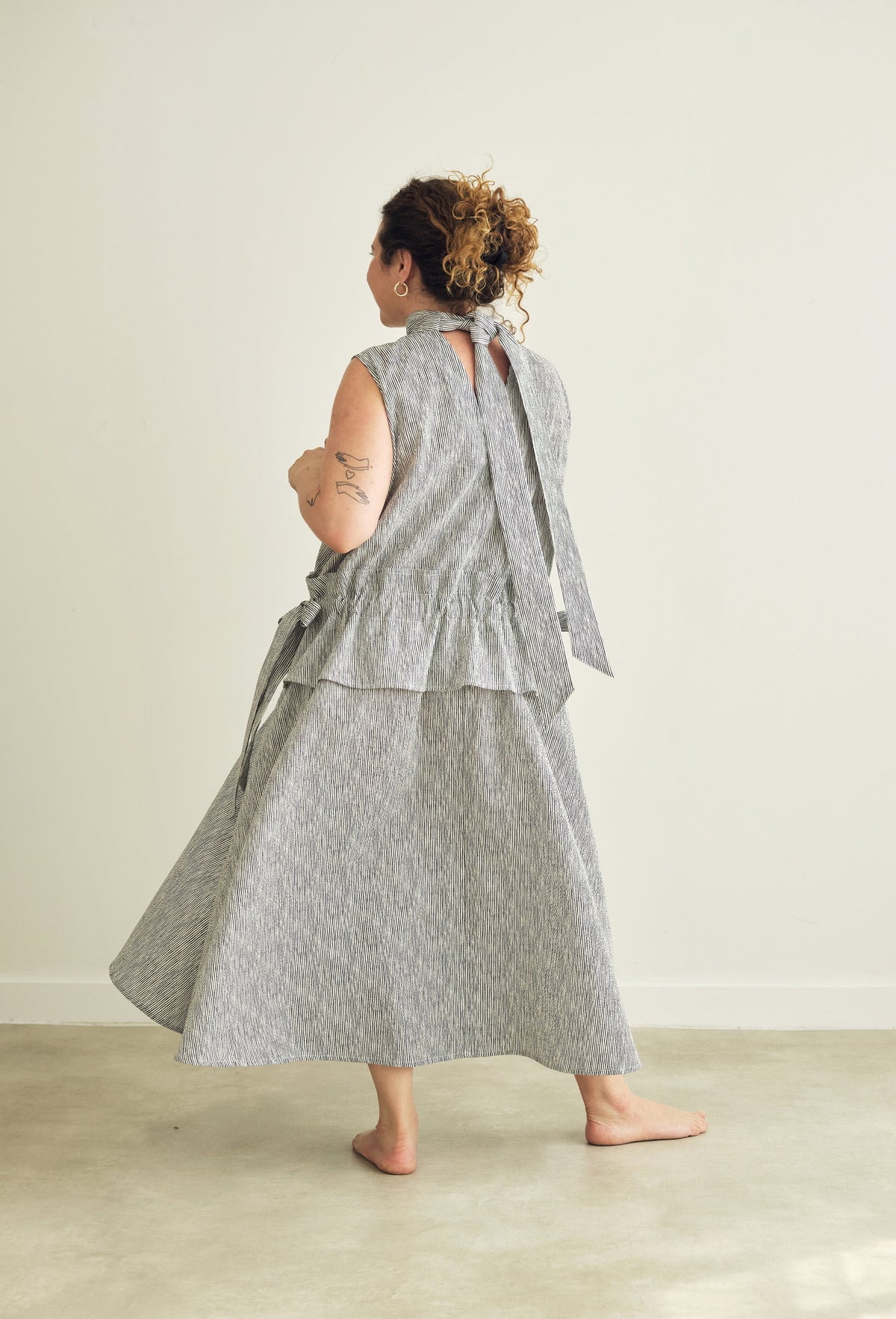 A woman wearing a sleeveless grey and white striped dress with a tie-back neckline, shown from behind in a minimalist studio setting.