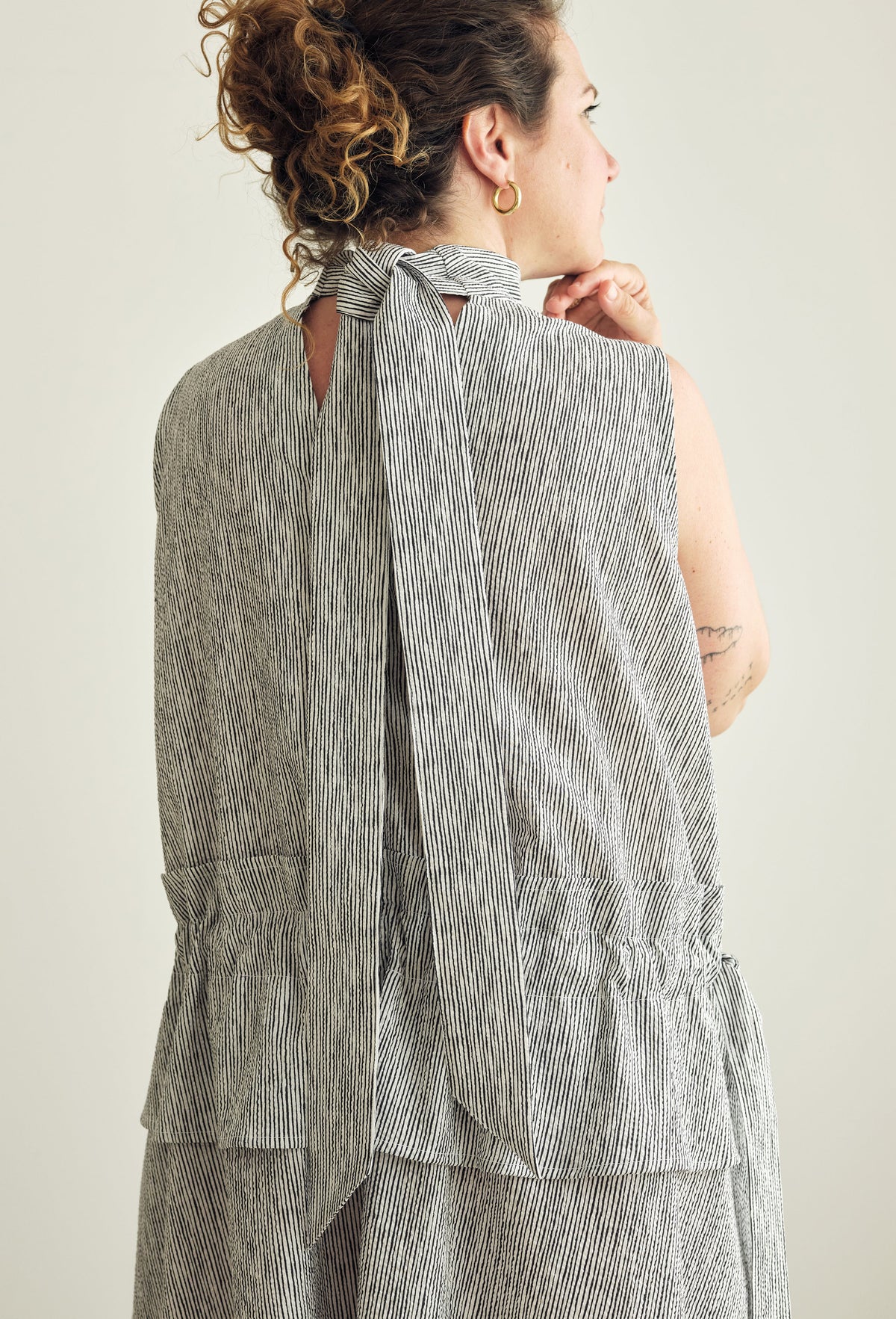 A woman shown from the back wearing a sleeveless grey and white striped dress with a tie-neck detail, photographed against a plain light background.