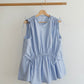 A light blue sleeveless top with tie details on the shoulders and waist, hanging on a wooden hanger from a wooden clothes rack against a plain off-white wall.