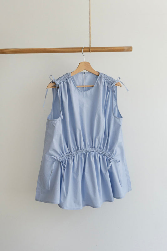 A light blue sleeveless top with tie details on the shoulders and waist, hanging on a wooden hanger from a wooden clothes rack against a plain off-white wall.