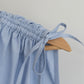 Close up of shoulder tie detail on light blue Paloma top on a wooden hanger against a plain light gray background.