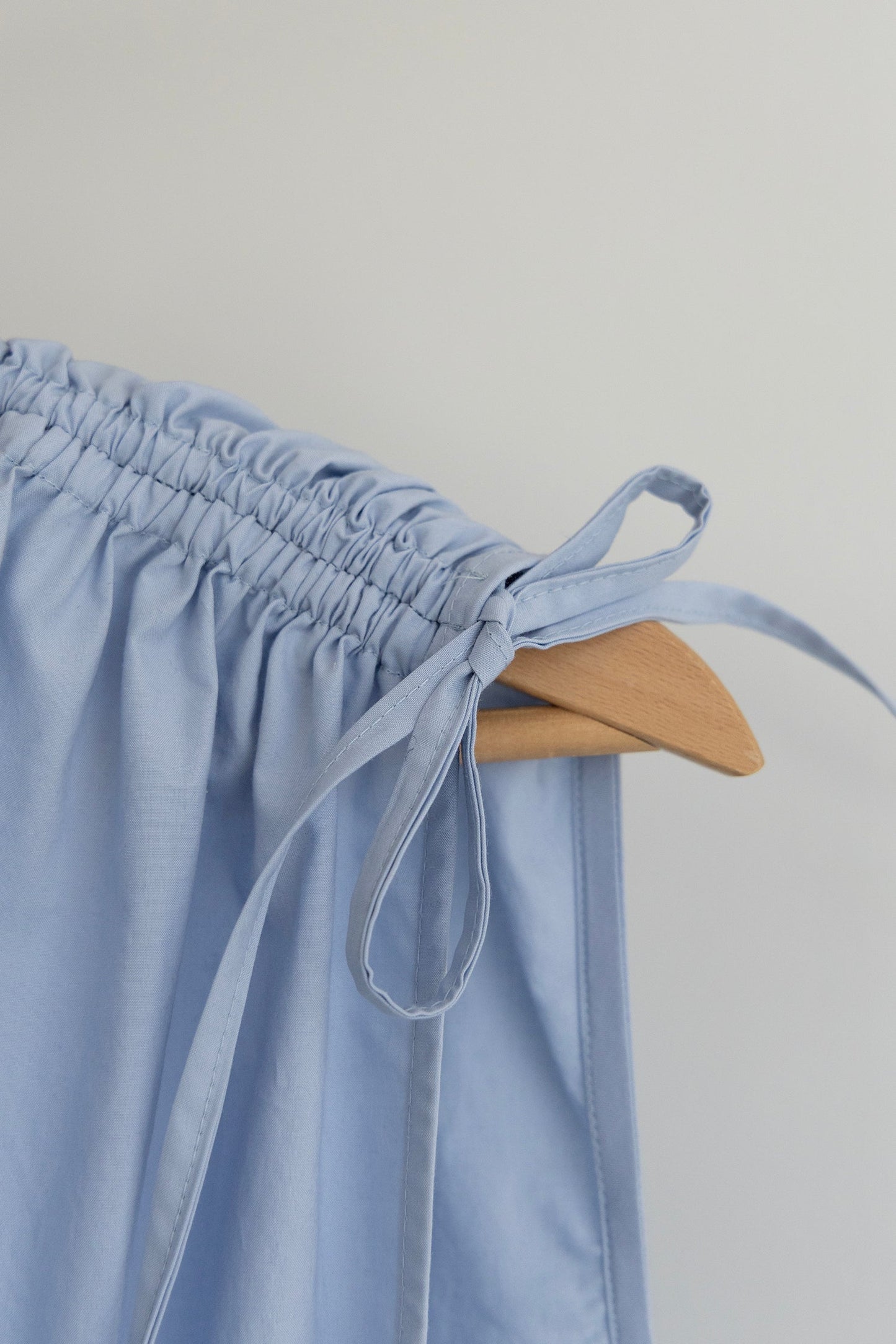 Close up of shoulder tie detail on light blue Paloma top on a wooden hanger against a plain light gray background.