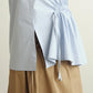 A close-up of a person wearing a light blue Paloma top with a gathered tie waist  detail and a tan skirt against a plain light beige background.