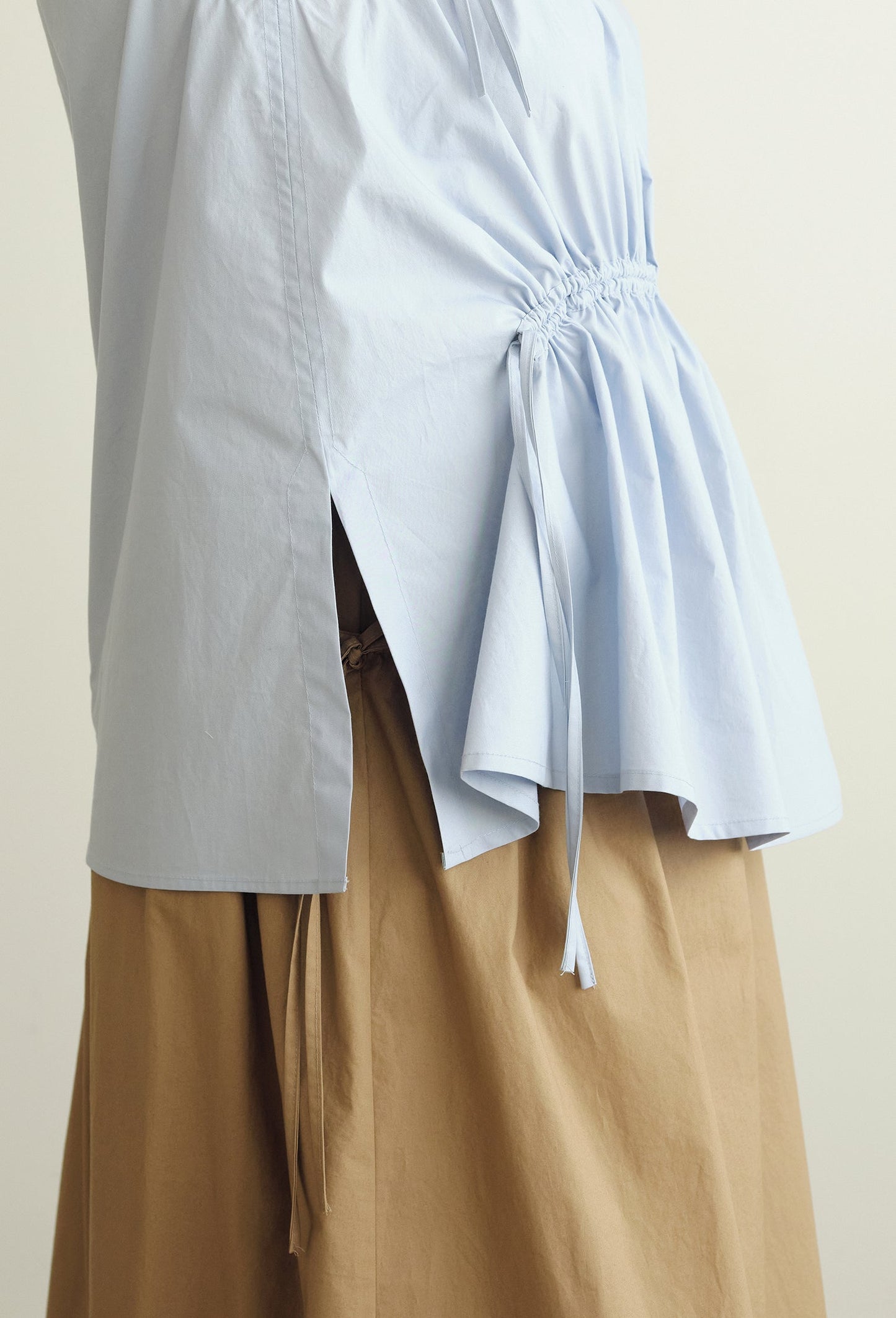 A close-up of a person wearing a light blue Paloma top with a gathered tie waist  detail and a tan skirt against a plain light beige background.