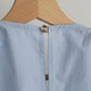 A light blue Paloma top hanging on a natural wood hanger, shown from the back with a central seam and a single button closure near the top.