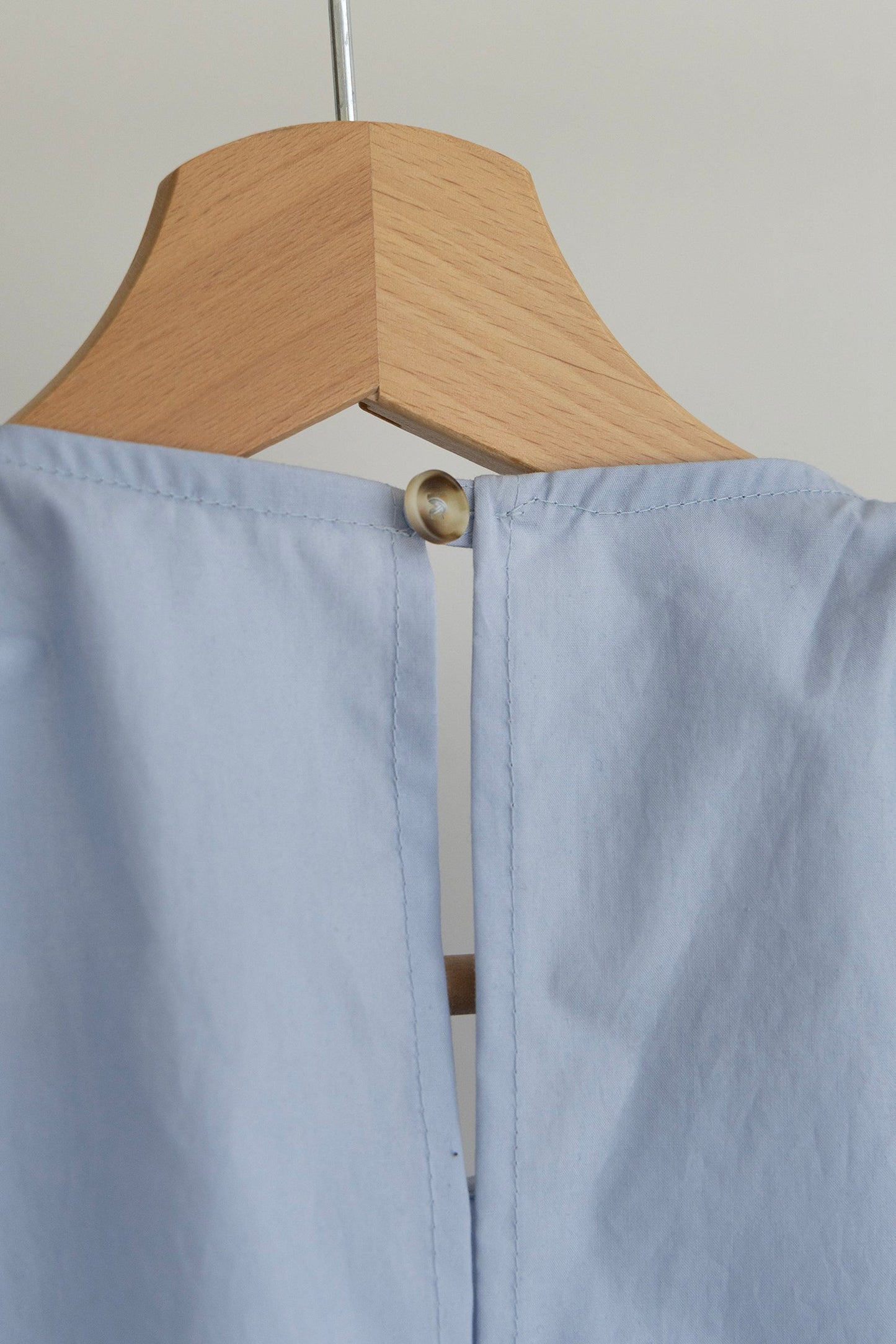 A light blue Paloma top hanging on a natural wood hanger, shown from the back with a central seam and a single button closure near the top.