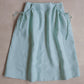 A light blue linen skirt with gathered drawstring pockets, laid flat on a textured beige background.