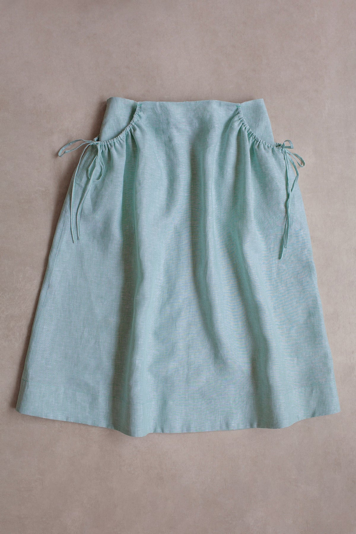 A light blue linen skirt with gathered drawstring pockets, laid flat on a textured beige background.