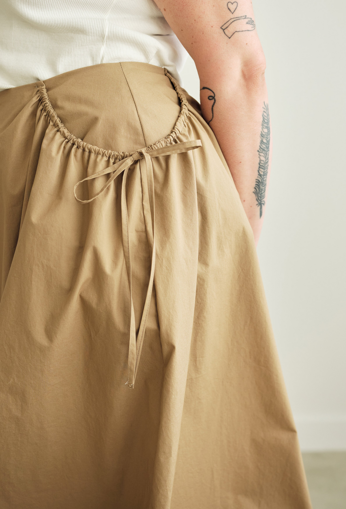 Close up of a person wearing a beige skirt with a white top, standing against a neutral background.