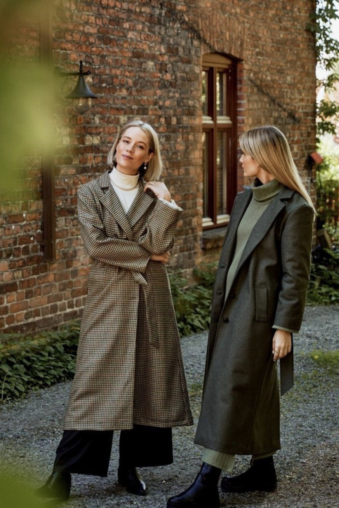 Two white women standing wearing long neutral coloured coats against a brick building