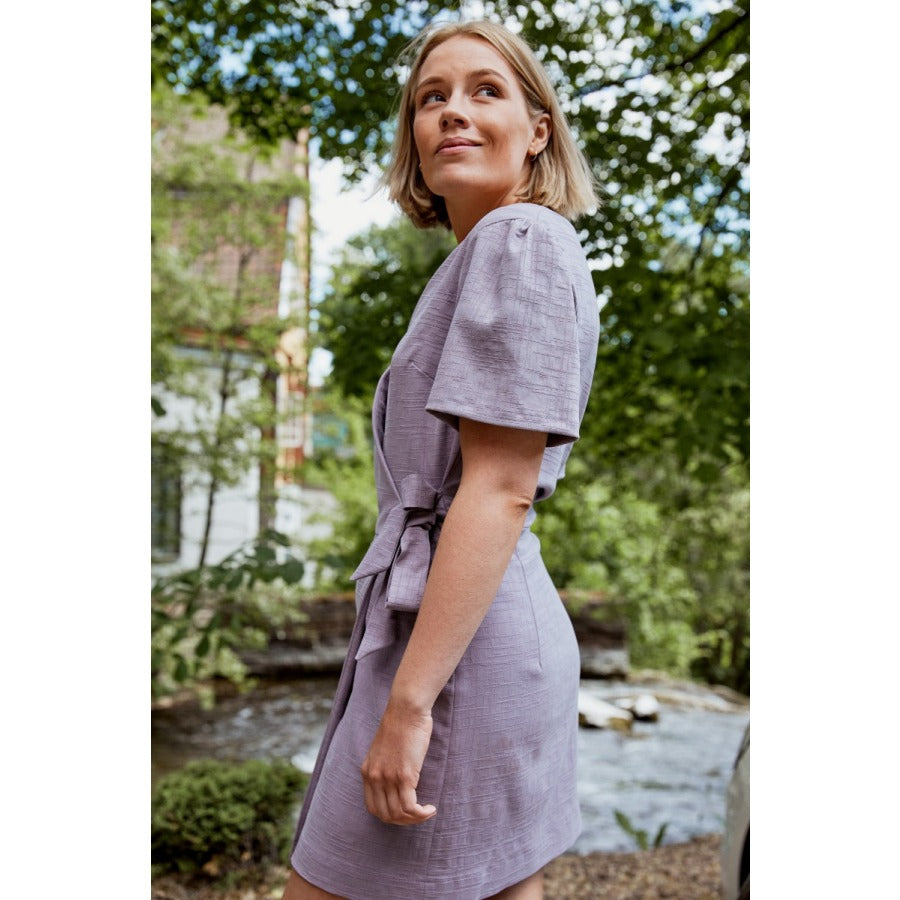 White woman with short blonde hair standing in a park wearing a light purple short sleeved wrap dress