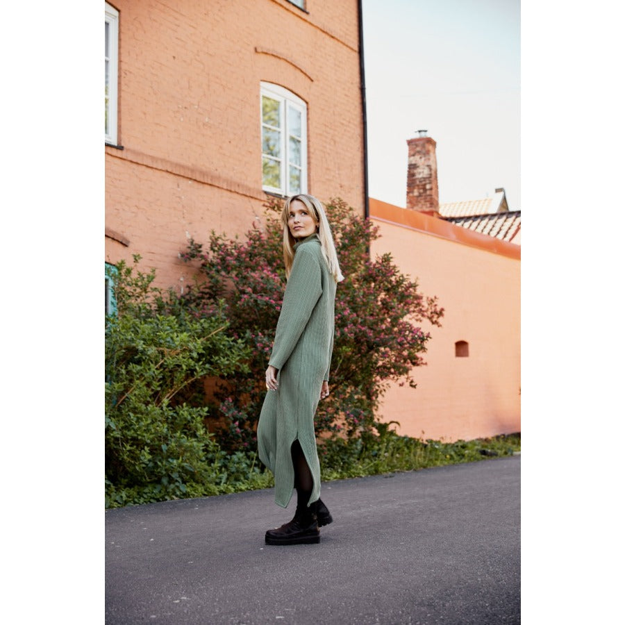 White woman with long blonde hair standing on a driveway in a long green sweater dress