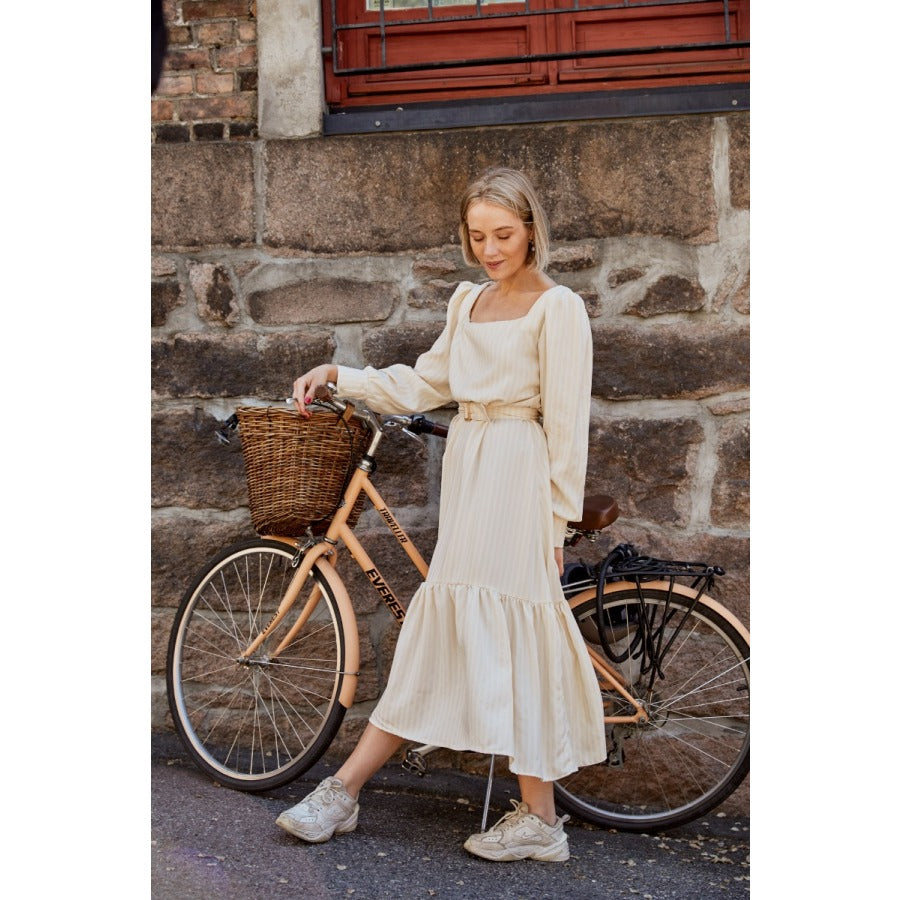 White woman with short blonde hair standing next to a Dutch bike wearing a long cream dress with  sneakers