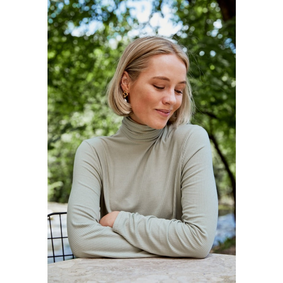 White woman with short blond hair sitting at a table with her arms crossed smiling and wearing a light green turtleneck