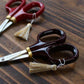 Embroidery scissors with red handles, brown handles and gold tassels sitting on top of a dark wooden background