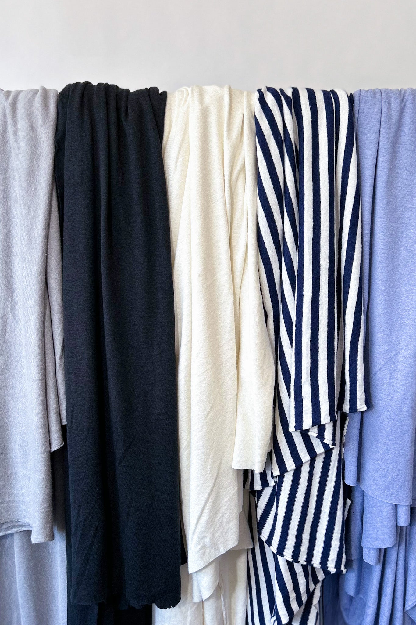 Row of hemp jersey fabrics hanging on rail in grey, black, natural white, navy/white stripe and periwinkle blue